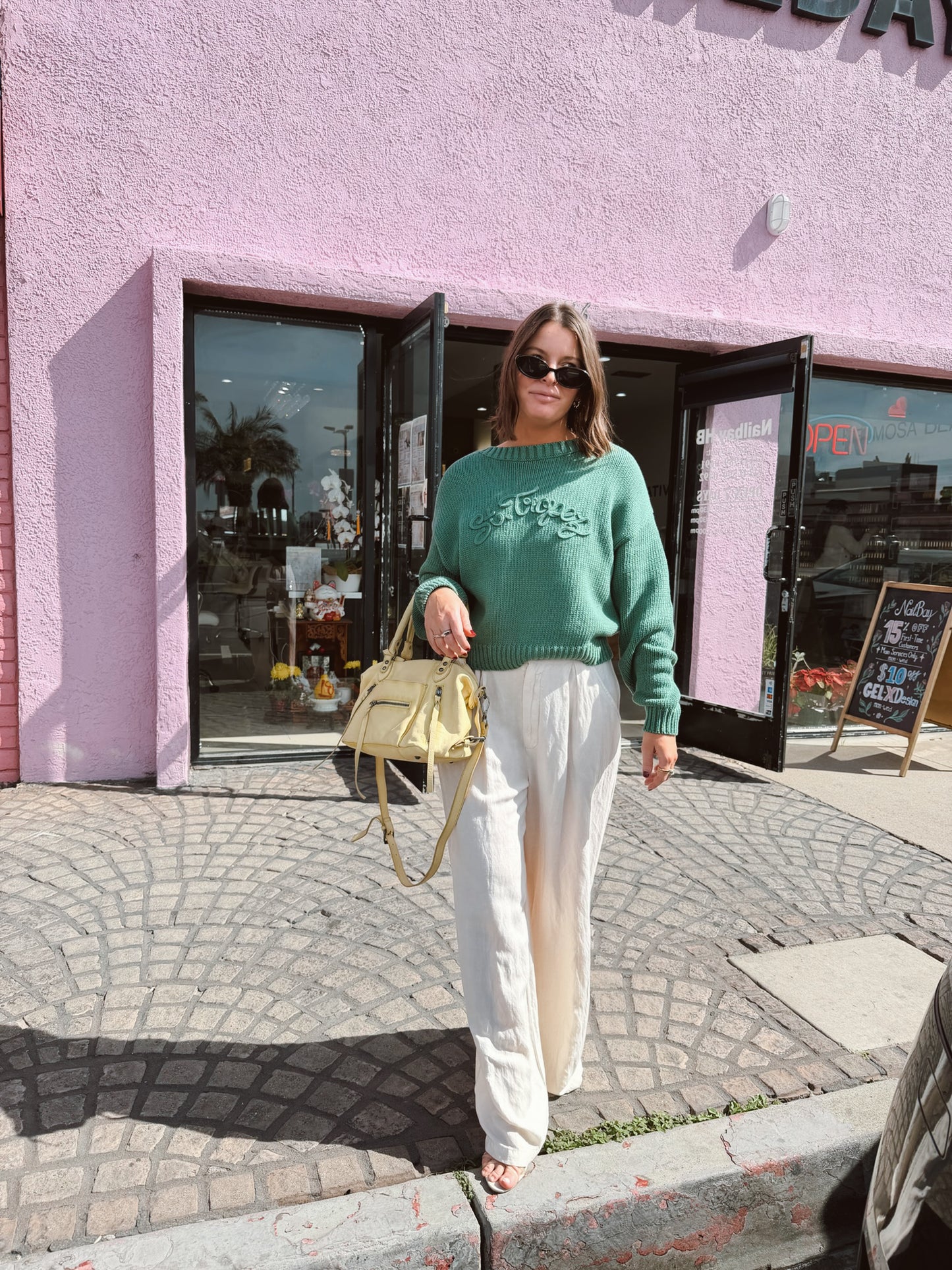 Woman in green crop boat neck sweater stands by pink building, carrying a yellow handbag. Sweater appears lightweight.