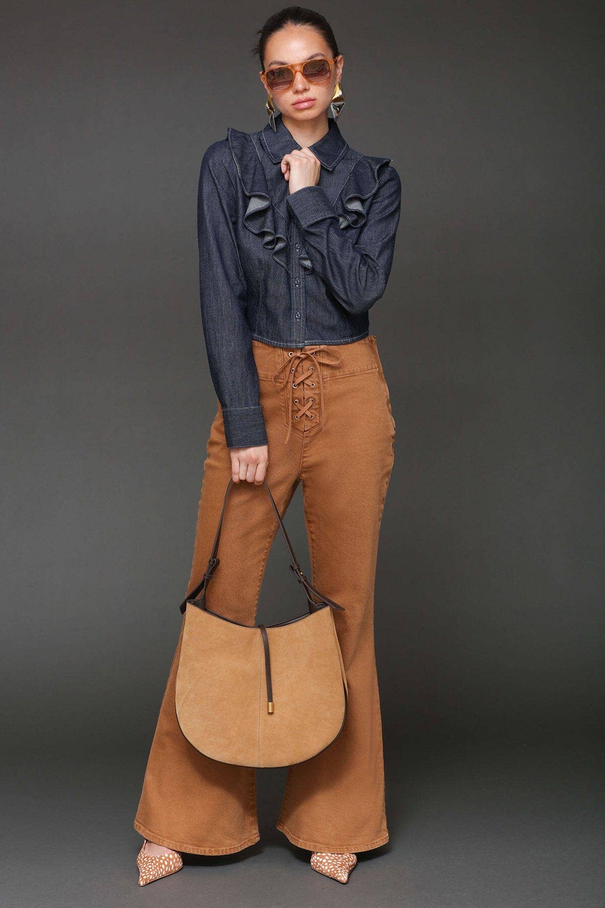 Woman in sunglasses wears a cropped denim shirt, tan flared pants, and carries a tan handbag against a gray backdrop.