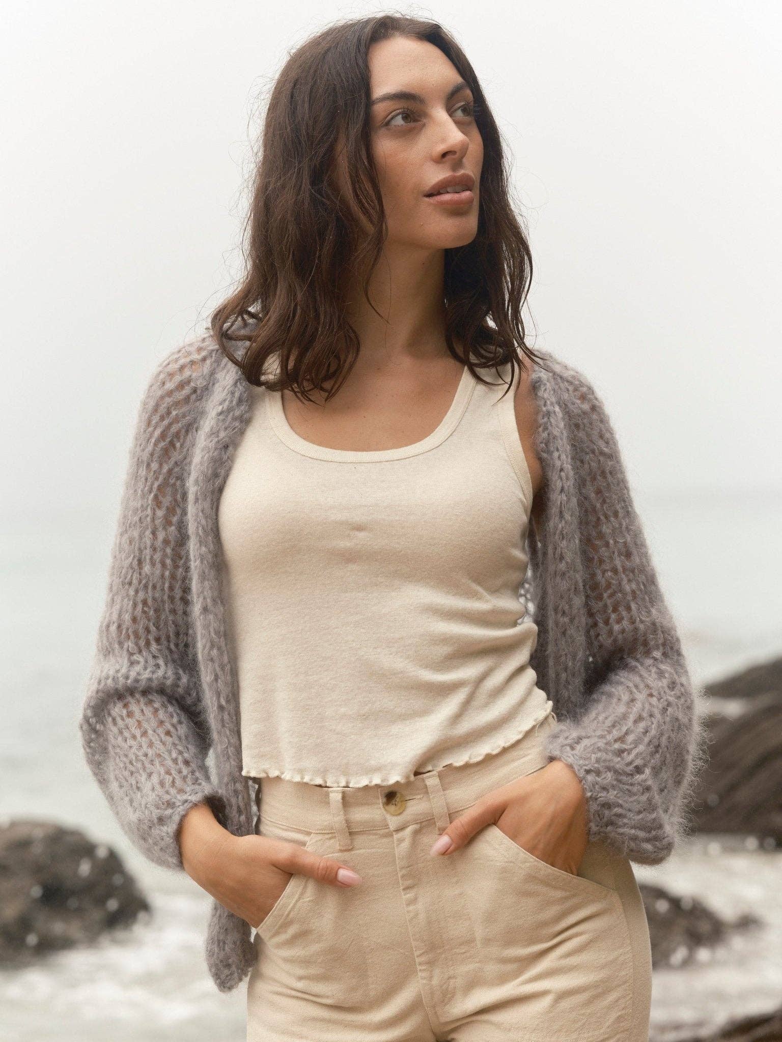 Woman in a cloud-patterned wool cardigan by a rocky shore on an overcast day, looking to the side.