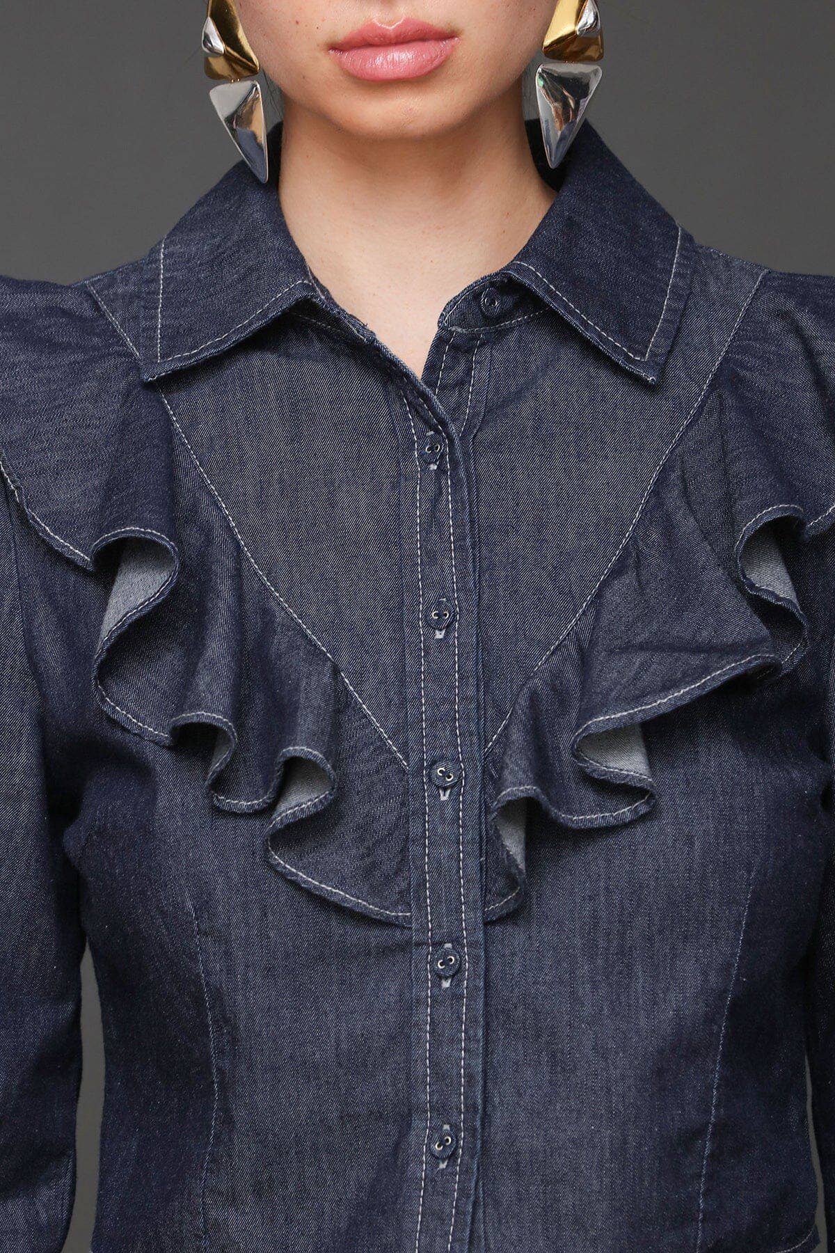 Woman in a blue denim cropped shirt with ruffles and large silver earrings, visible from shoulders up.