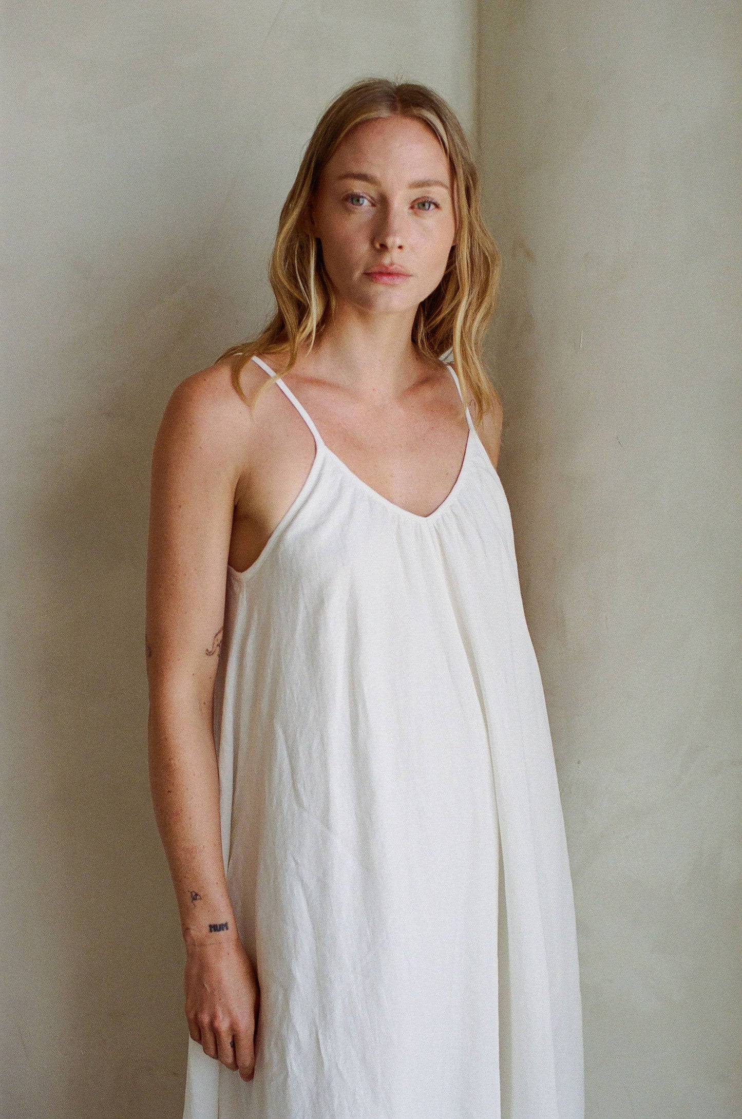 Woman with long blonde hair wears a white linen maxi dress, standing by a light wall and facing the camera.