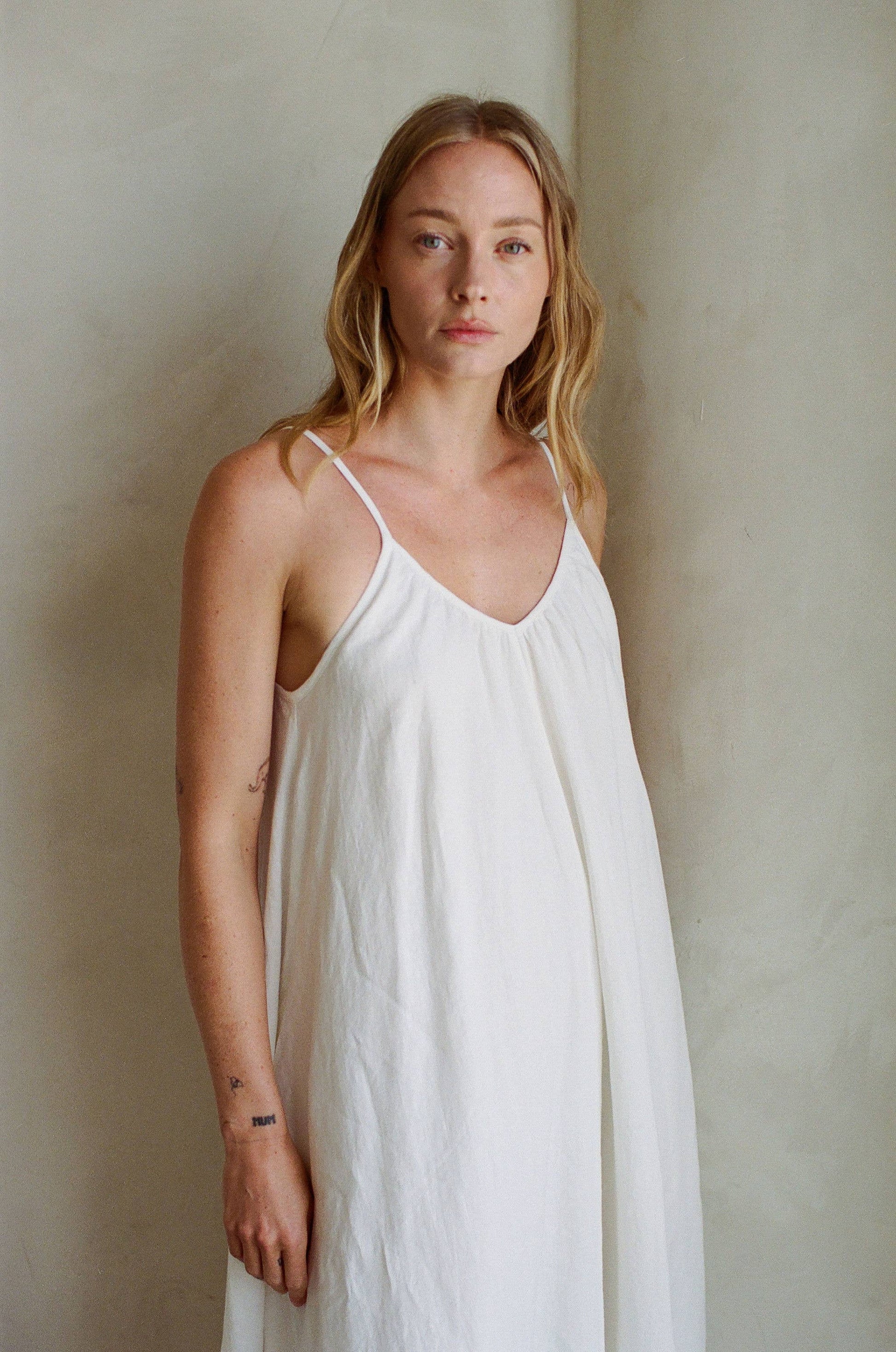 Woman with long blonde hair wears a white linen maxi dress, standing by a light wall and facing the camera.