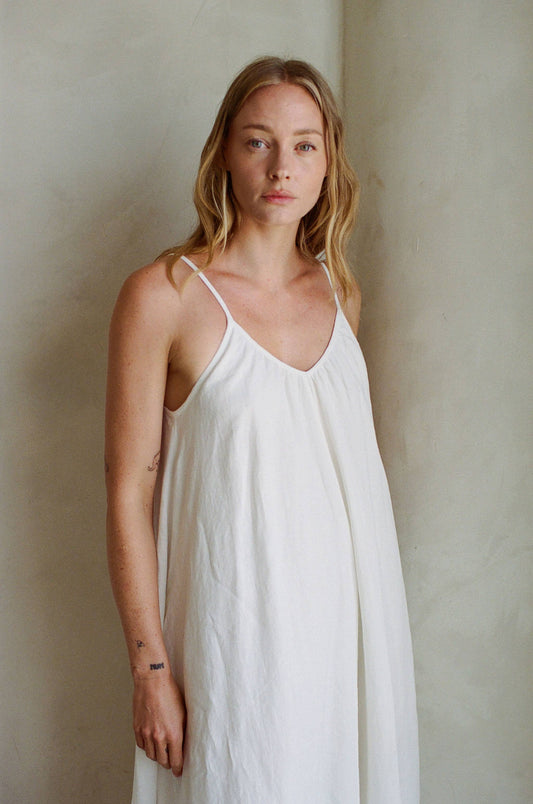 Woman with long blonde hair wears a white linen maxi dress, standing by a light wall and facing the camera.