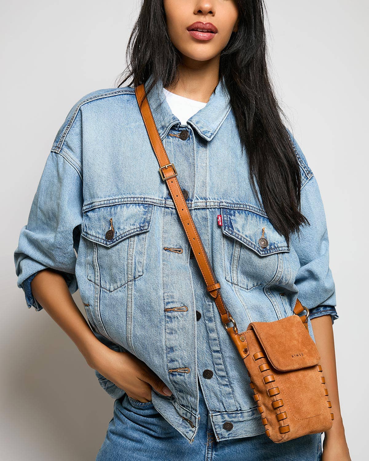 Woman in denim jacket with a leather crossbody phone bag in neutral tones, worn over shoulder against plain backdrop.