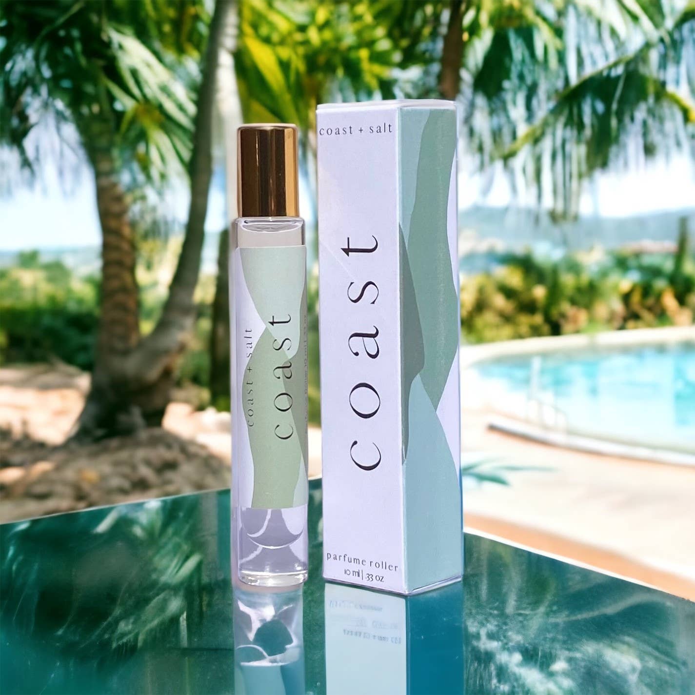 Clear glass perfume roller and box on a glass table near a pool, with palm trees visible in the background.