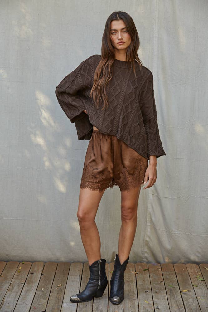 Woman wears brown knit sweater, black ankle boots, and tan shorts on a wooden floor.