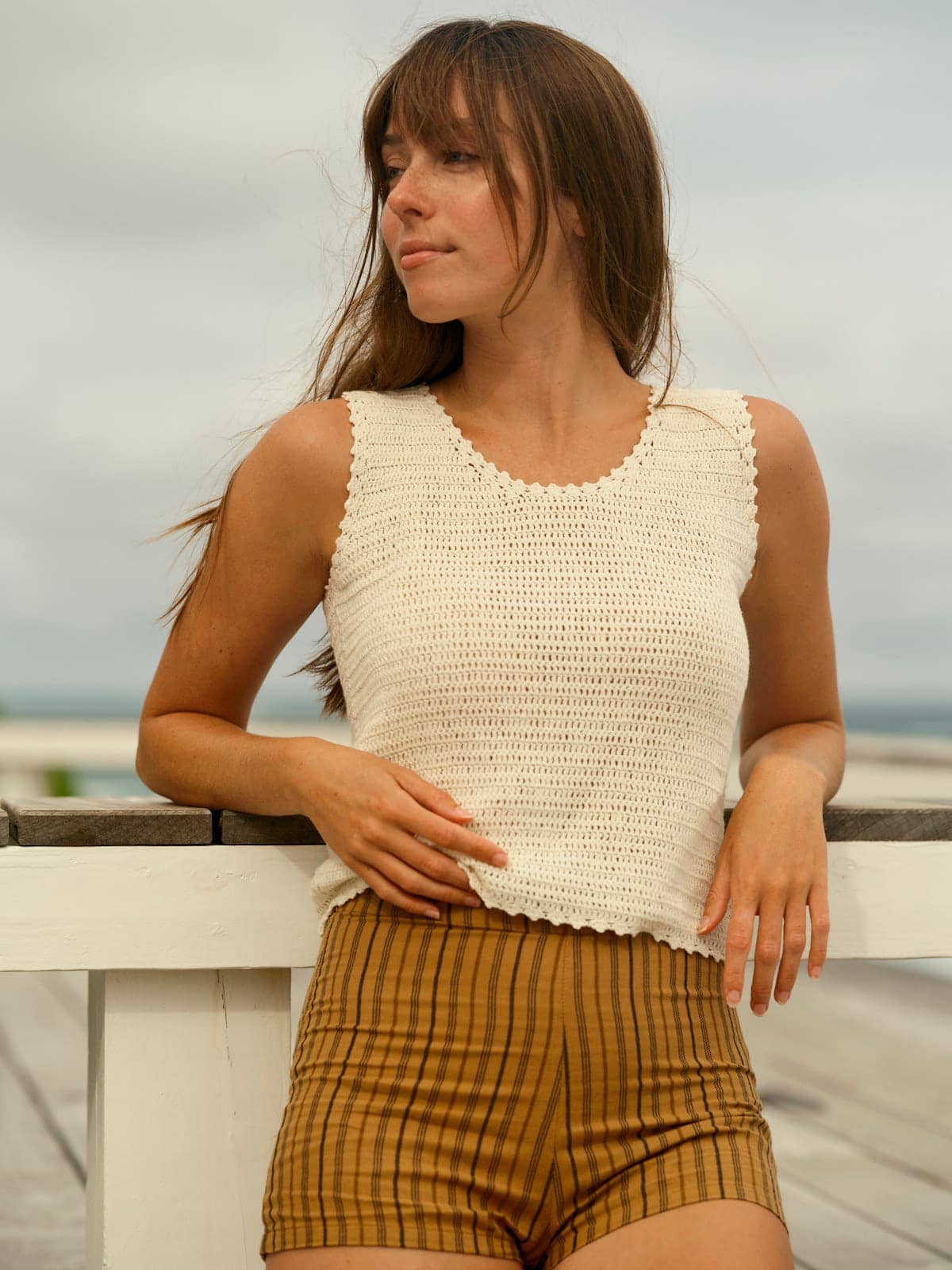 Woman in crochet tank top and brown striped shorts leans on a white outdoor wooden railing.
