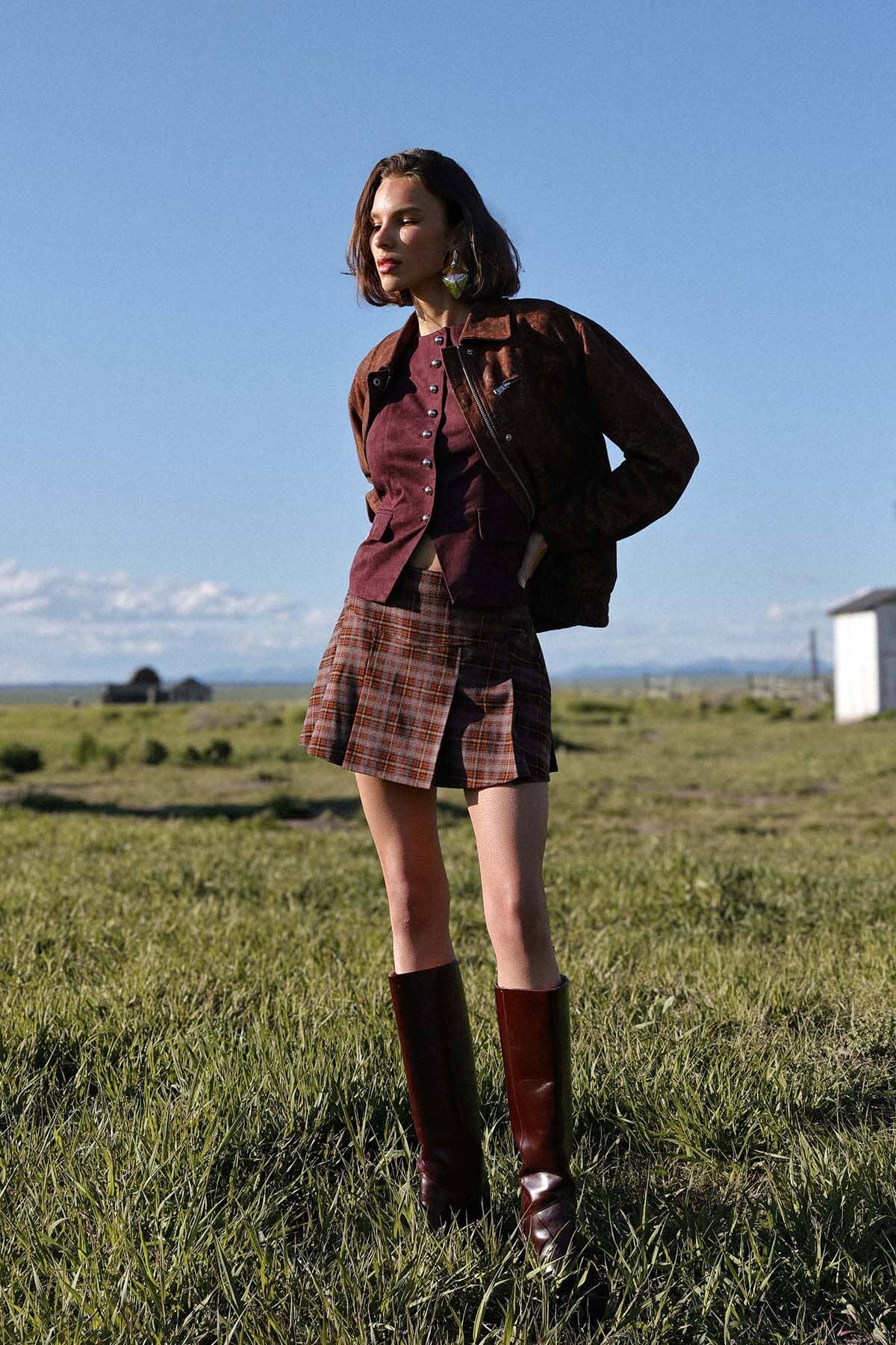 Woman on grass wearing plaid skirt, burgundy top, maroon boots, and a floral print relaxed-fit bomber jacket.