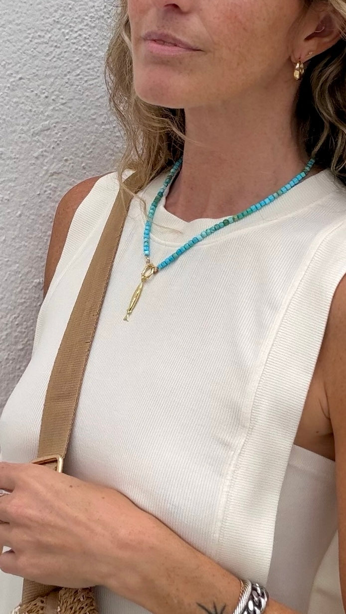 Woman in parchment open-side top, turquoise bead necklace, gold earrings, and tan shoulder bag strap.