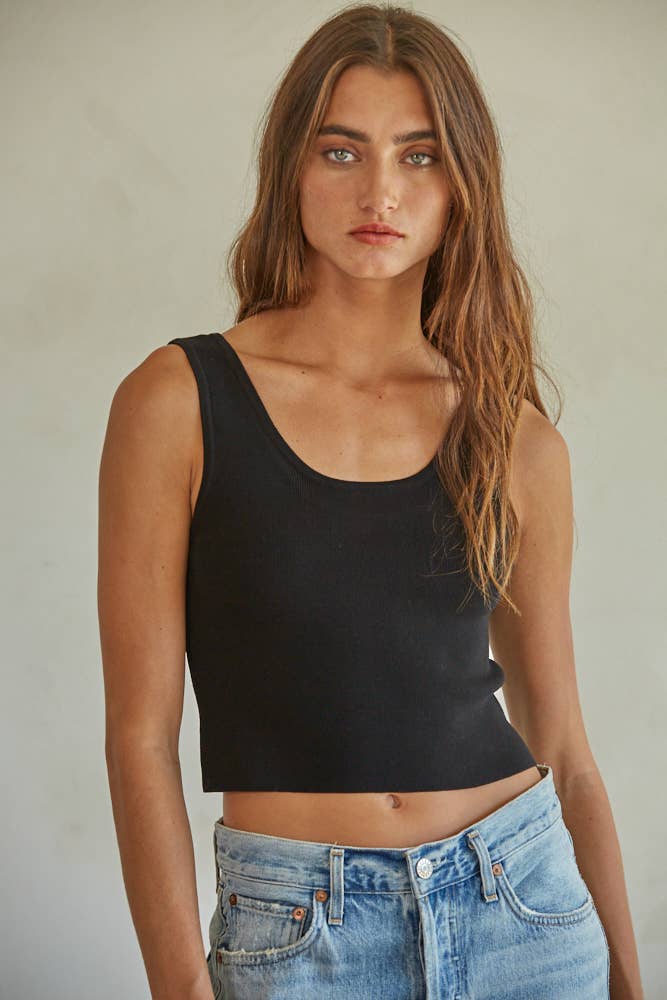 Woman with long brown hair wears a black ribbed tank top and blue jeans, standing against a neutral background.