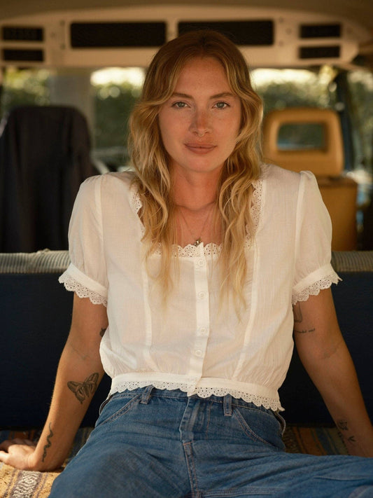 Woman with wavy blonde hair wears a light linen top and denim jeans, seated in a van, facing the camera.