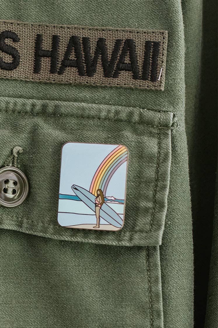 Rainbow surfer enamel pin in vibrant colors on a green shirt, positioned beneath a Hawaii patch.