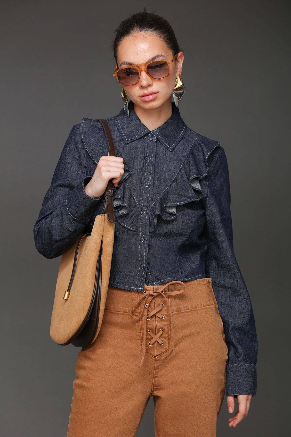 Woman in sunglasses wears a cropped denim shirt, tan lace-up pants, and carries a tan leather shoulder bag.