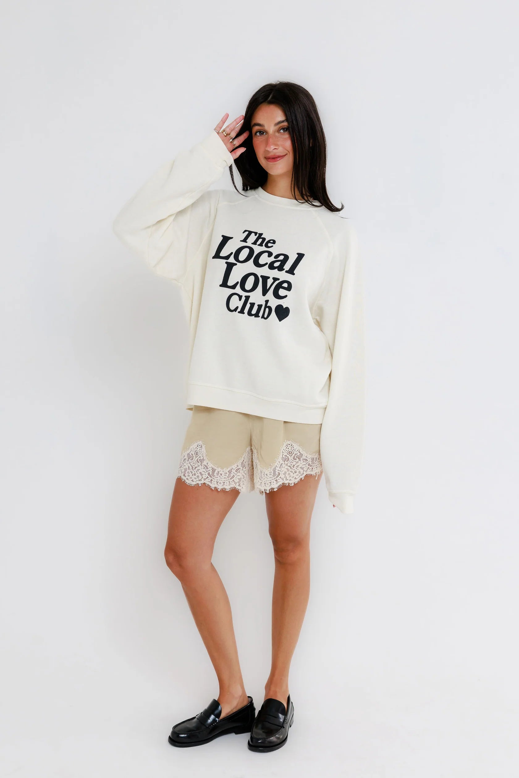 Woman wearing a white raglan sweatshirt, lace-trim shorts, and black loafers, standing against a plain white background.