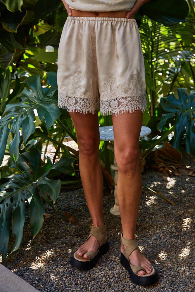 Person wearing ivory cotton shorts stands on gravel, surrounded by dense green foliage.