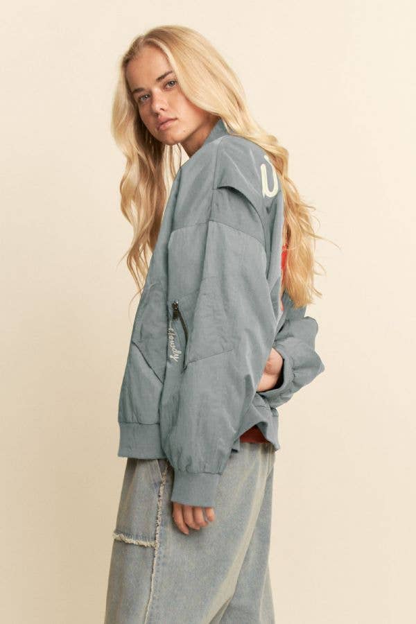 Woman with long blonde hair wearing a dark embroidered bomber jacket and loose denim jeans, suitable for casual wear.
