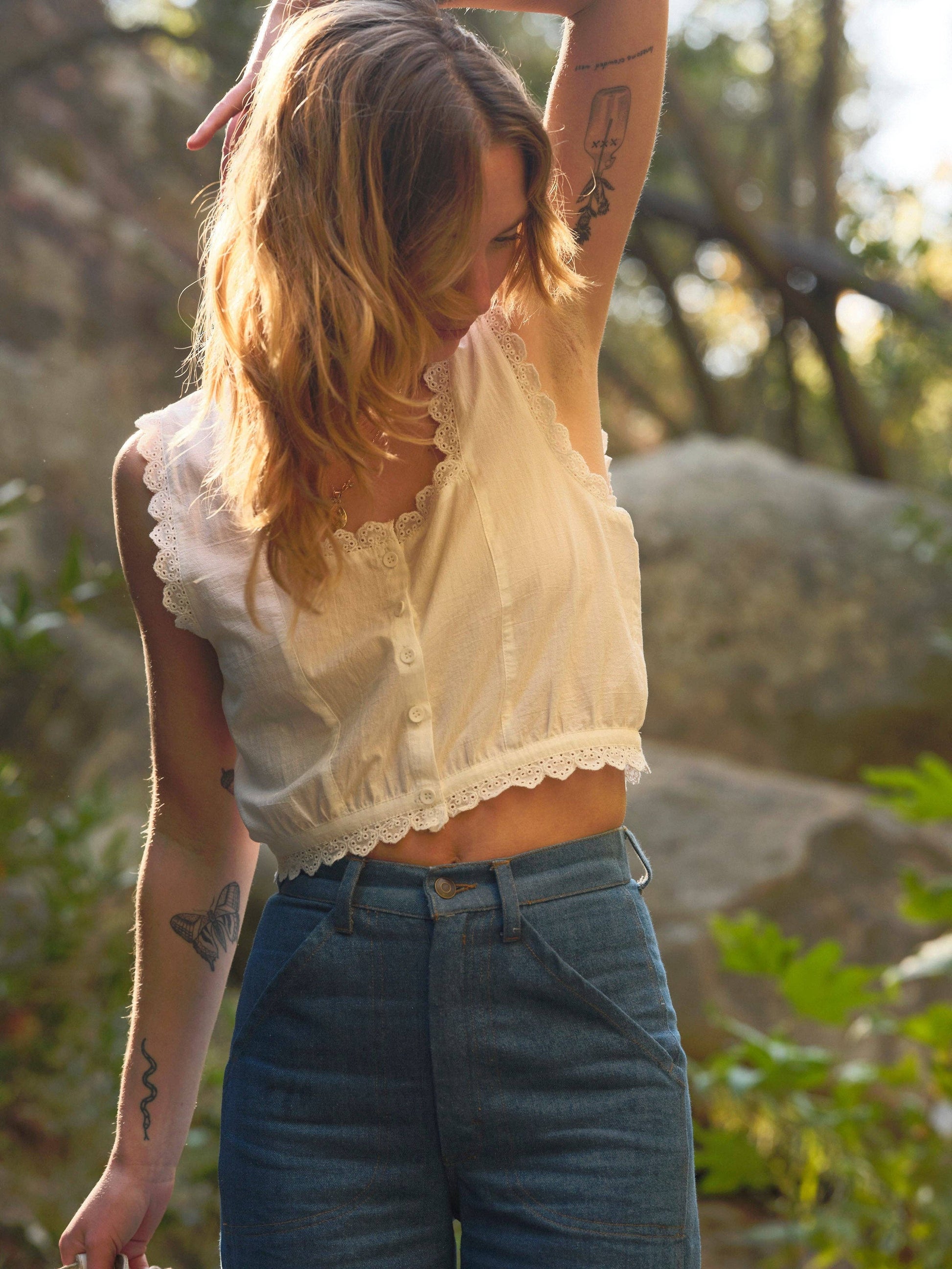 Woman in a patterned cotton top and blue jeans stands outside with one arm raised, looking downward.