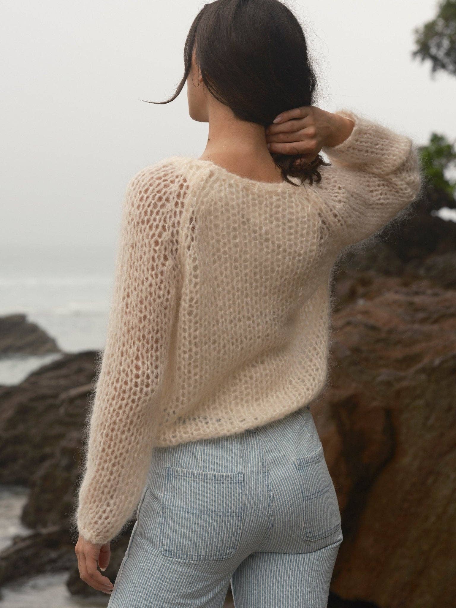 A person in a gray knit cloud sweater stands by rocky shore, facing the ocean.