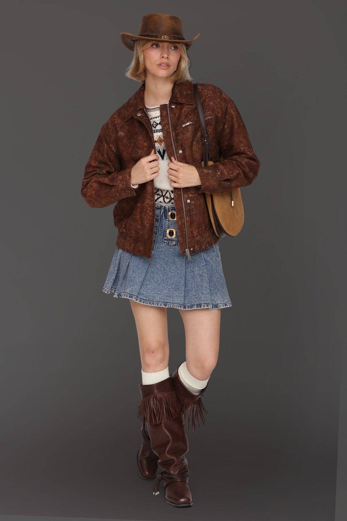 Woman in a floral print bomber jacket, blue denim skirt, tan cowboy hat, and brown suede fringed boots.
