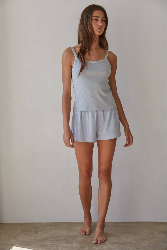 Woman wears a soft, light-colored cami and matching shorts set, standing barefoot against a plain white wall.