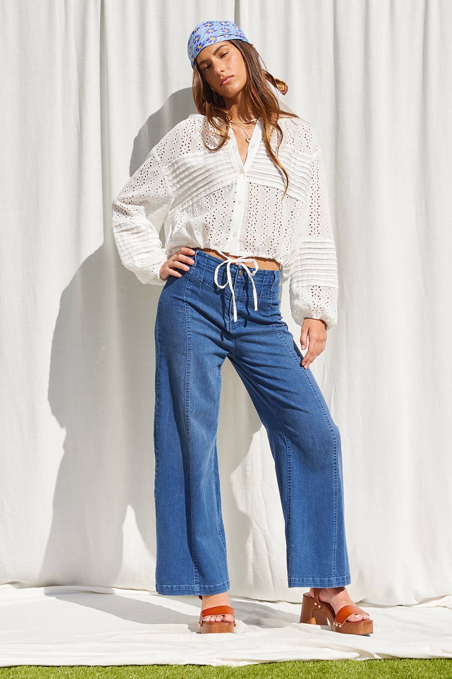 Woman in off-white top, blue wide-leg jeans, sandals, and bandana stands in front of a curtain.
