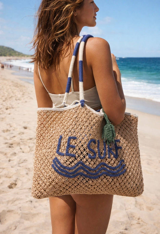 Woman on a beach in a swimsuit holds a macrame tote bag with blue lettering.