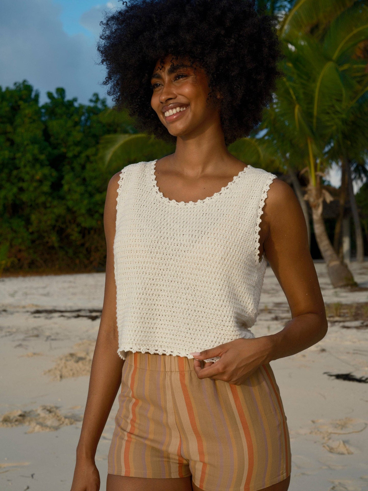 Woman in a white crochet tank top on a sandy beach with palm trees, showing breathable material ideal for warm weather.