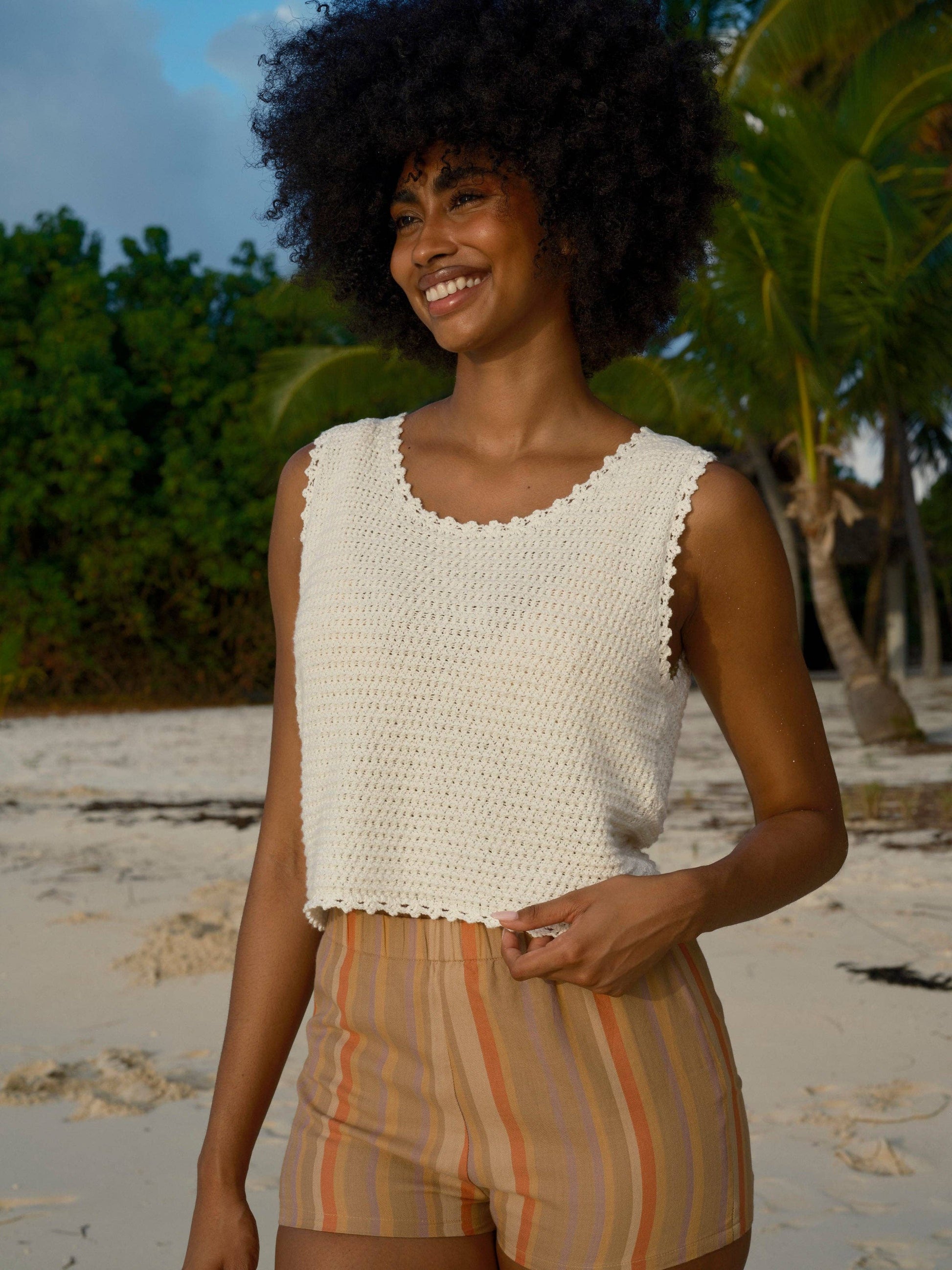 Woman in a white crochet tank top on a sandy beach with palm trees, showing breathable material ideal for warm weather.