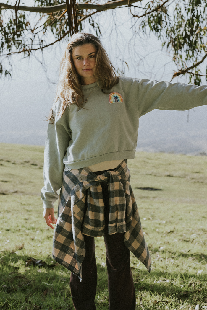Woman wearing eucalyptus green cropped sweatshirt and plaid shirt at waist, standing on grass.