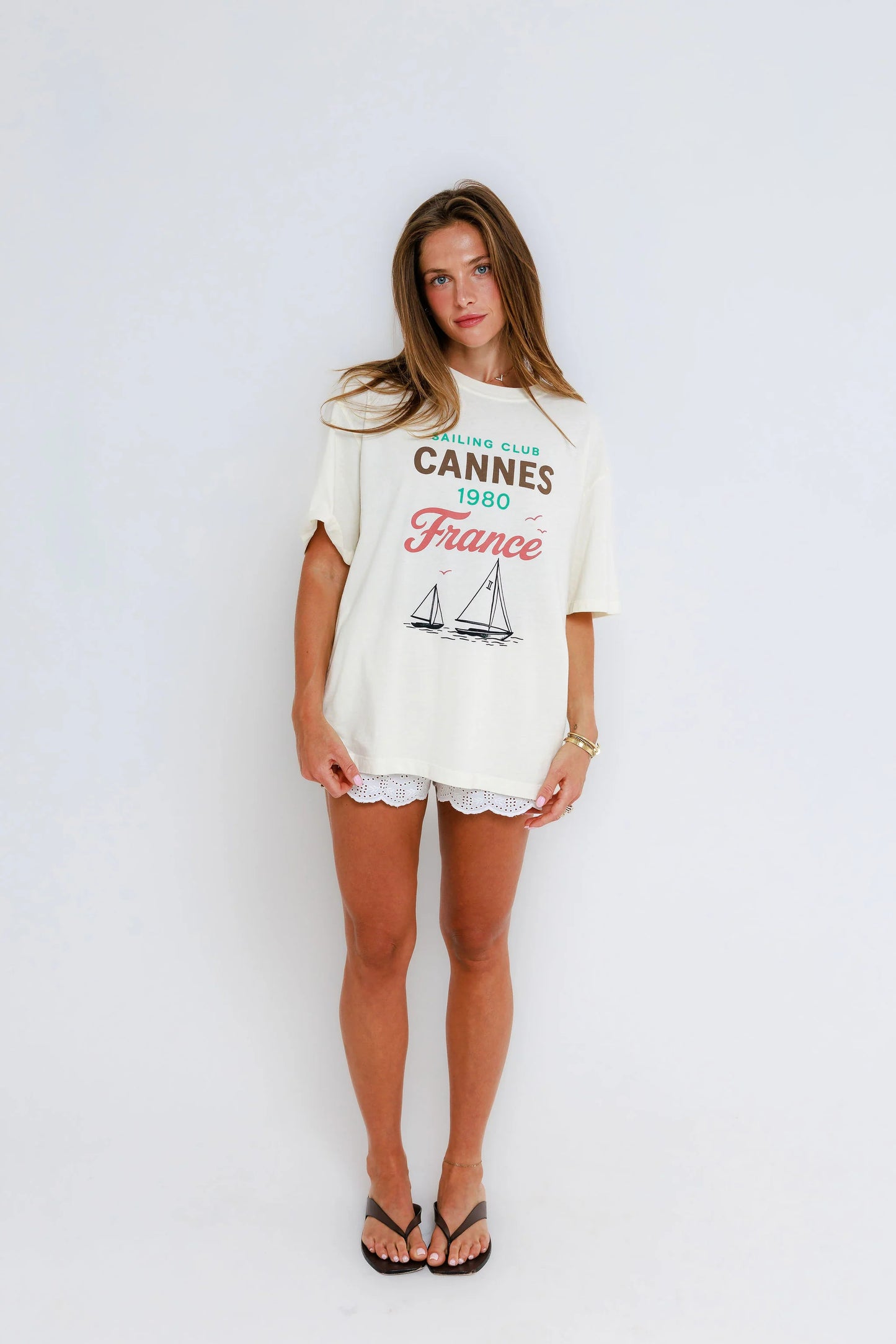 Woman in off-white cotton graphic tee, white shorts, and black sandals stands against a plain white background.