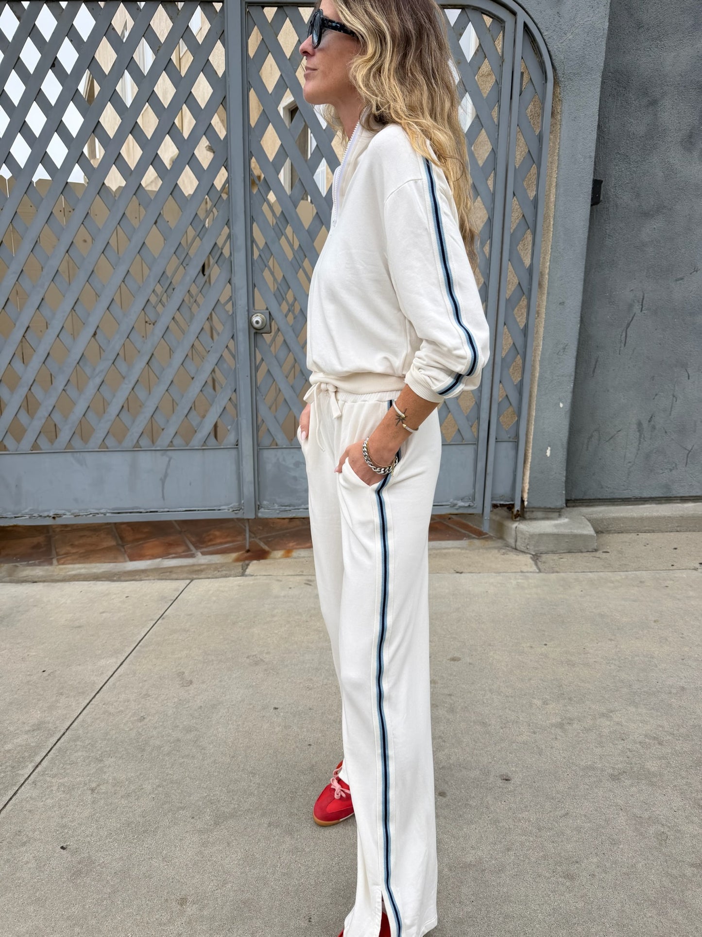 Woman in cream half-zip sweatshirt with blue stripes and red sneakers stands by gray gate, facing left.