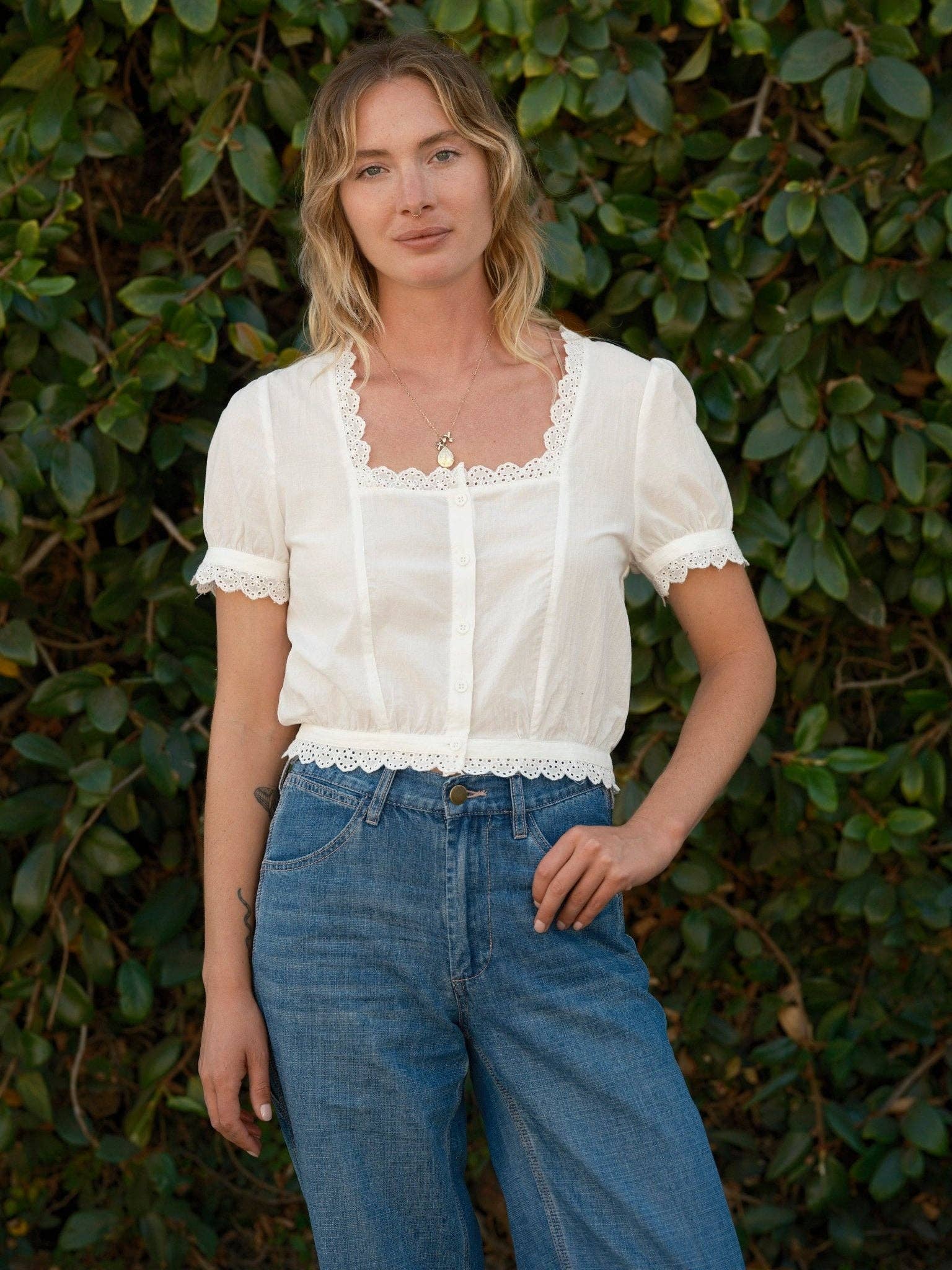 Woman in a cotton top and blue jeans stands facing the camera, green leafy bushes in the background.