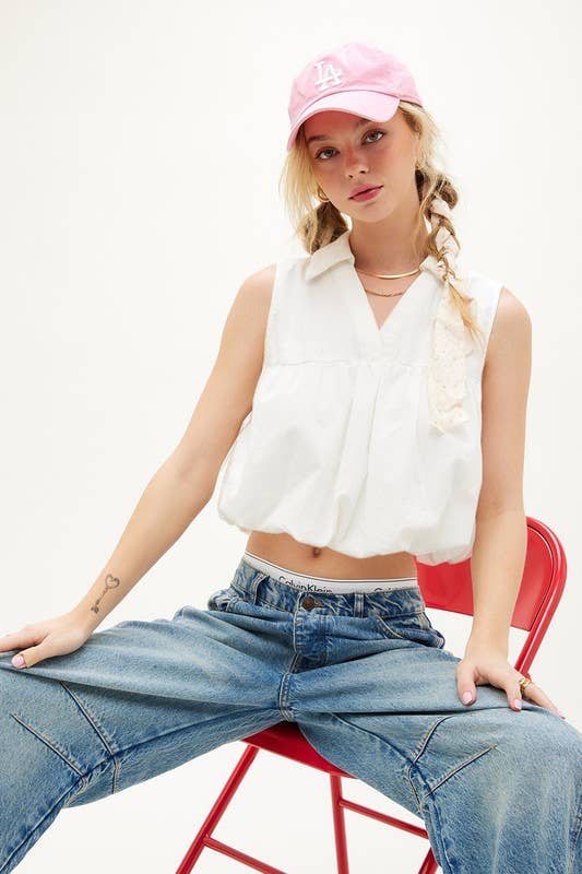 Woman wears off-white bubble top, pink cap, and jeans, seated on a red chair against a plain white background.