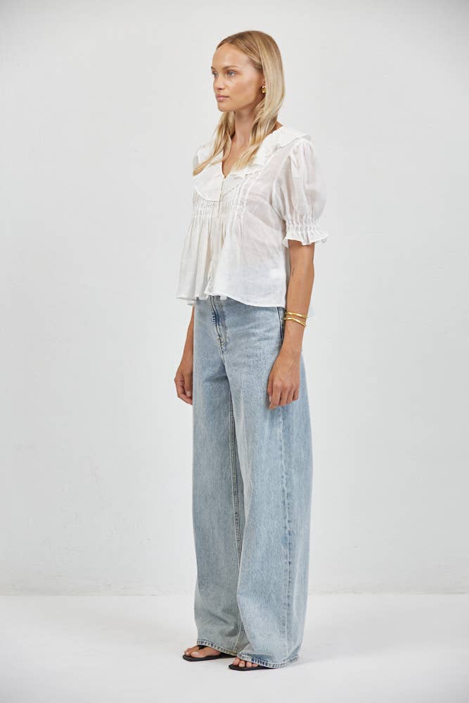 Woman wears a linen ruffle V-neck blouse and wide-leg light blue jeans, standing against a plain white background.