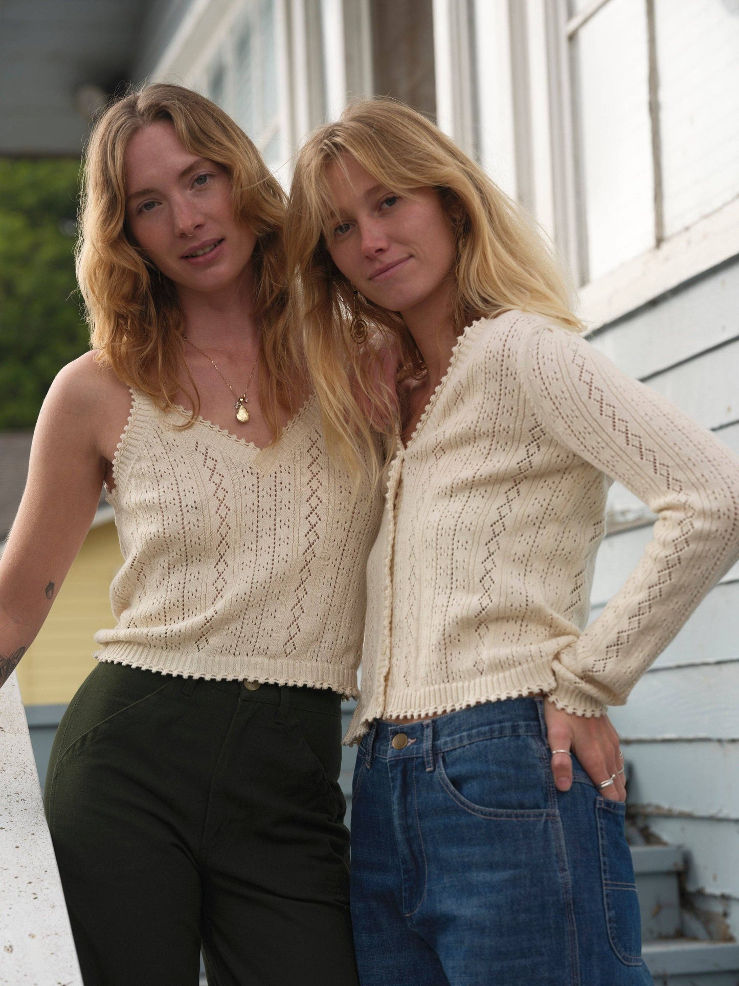 Two women wearing wool knit cardigans in muted tones, standing together outside a light blue wooden building.