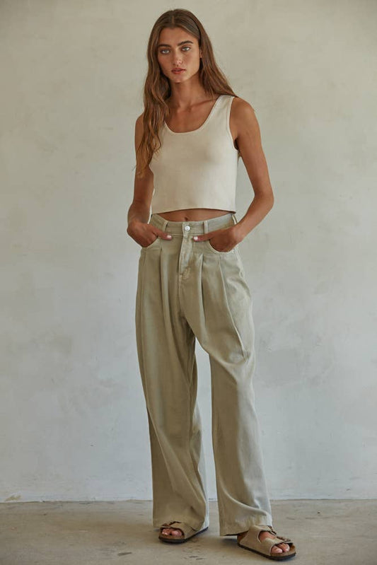 Woman with long hair in a sleeveless ribbed tank, loose beige cotton pants, and sandals stands by a plain wall.