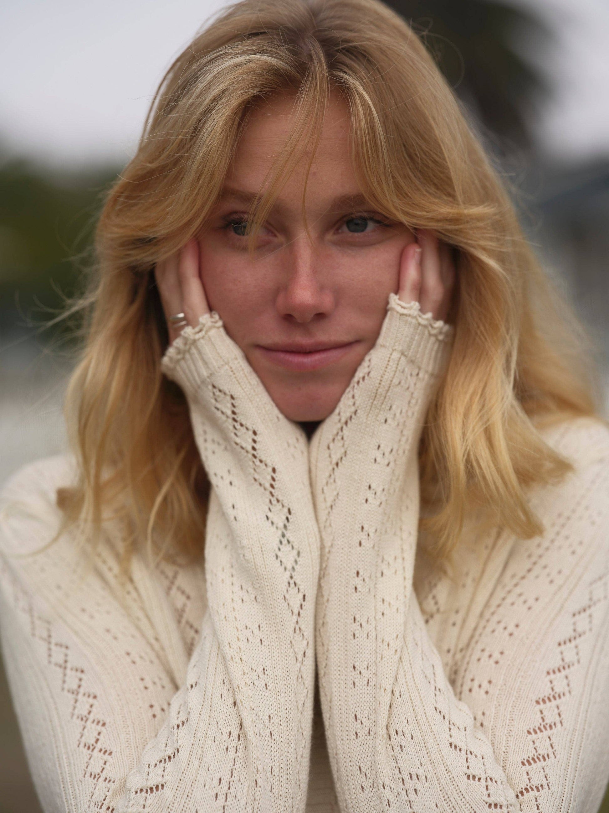 Woman wears a soft knit cardigan, light beige color, hands on face, standing outside.