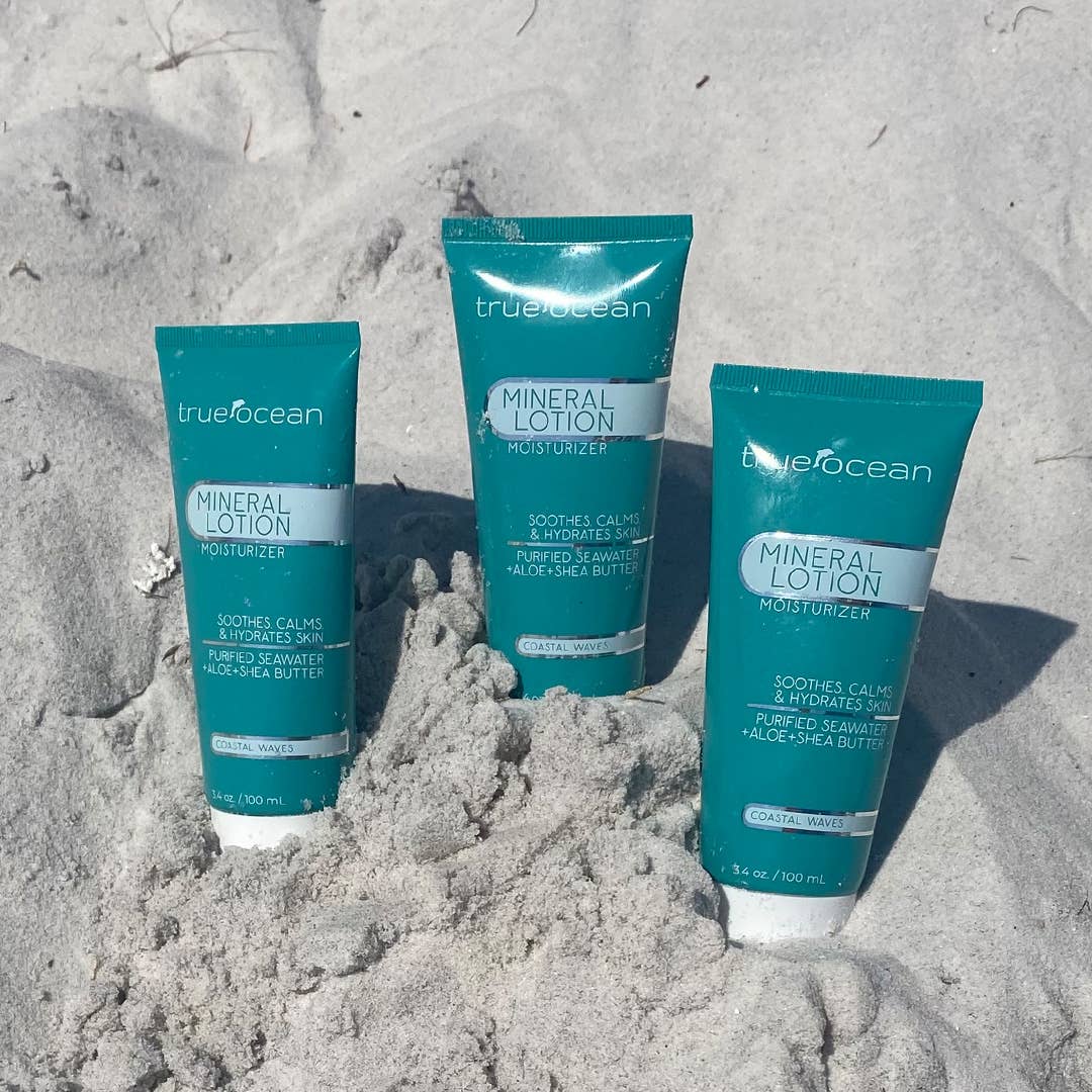 Three teal tubes of mineral lotion with shea butter stand upright in beach sand, intended for body care.