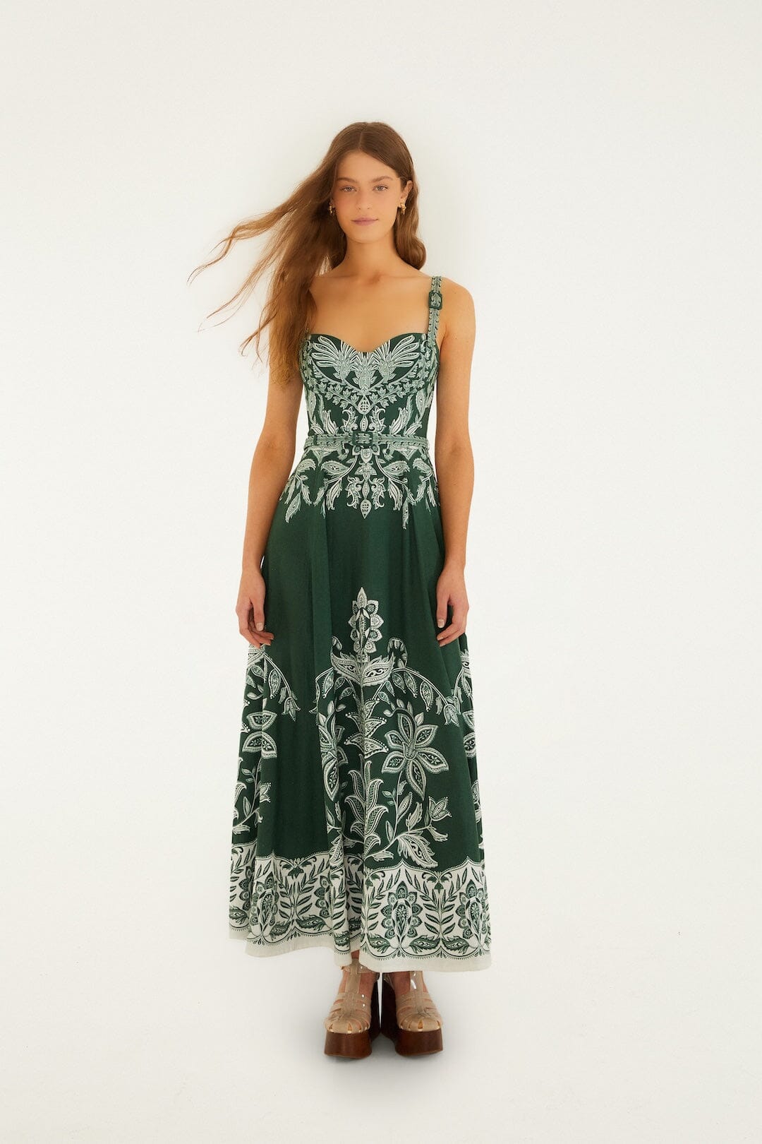 Woman in a dark green maxi dress with belt buckles and brown platform shoes, standing against a plain background.