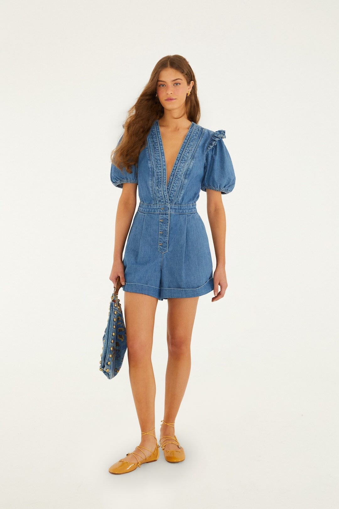 Woman wears a blue denim ruffle romper, carries a blue bag, and has yellow flats, standing in front of a white background.