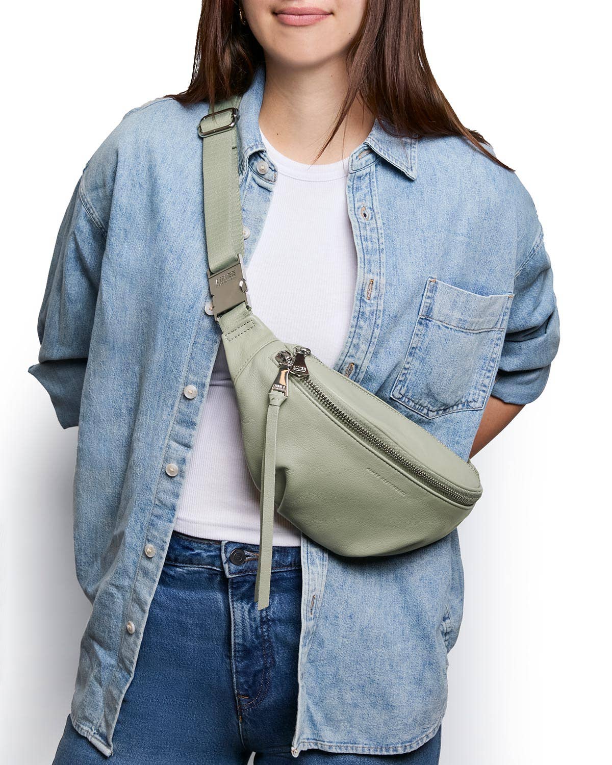 Woman wearing a light denim shirt and jeans with a green crossbody sling bag made from smooth material.