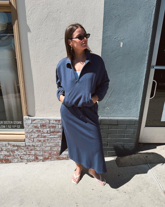 Woman wears a navy half-zip cotton sweatshirt, sunglasses, and flip-flops while standing on a sunlit sidewalk.