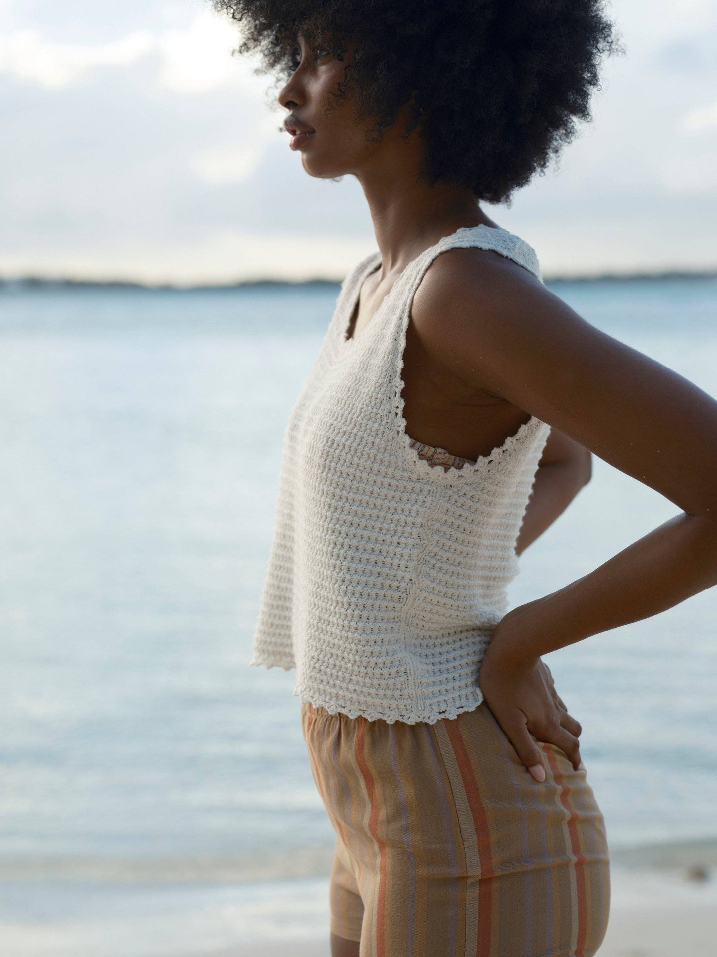 Woman in a cream crochet beach tank top stands by the water, hands on hips, facing left.