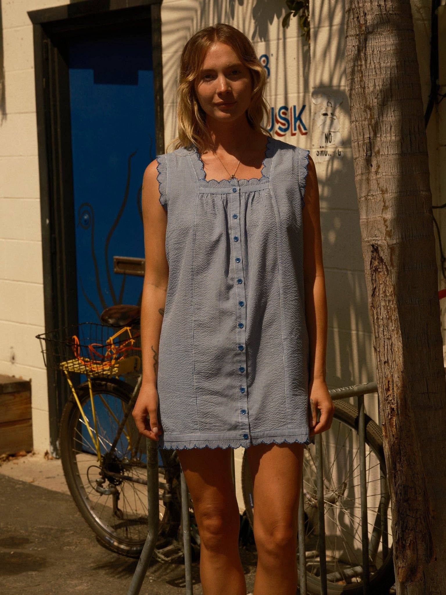Woman wearing a linen mini dress stands by a bike; sunlight creates sharp shadows on the wall and pavement.
