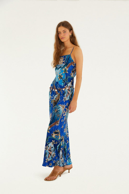 Woman in a blue maxi dress with heels stands against a white background. The dress is flowy and ankle-length.