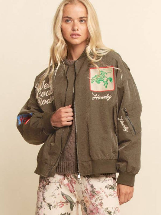 Woman with long blonde hair models an embroidered bomber jacket against a beige background.