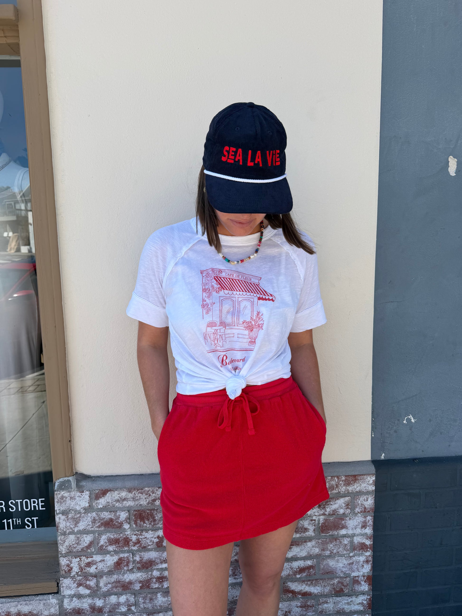 Woman in a white cap, white knotted shirt, and vermillion terry mini skirt stands by a brick wall, looking down.