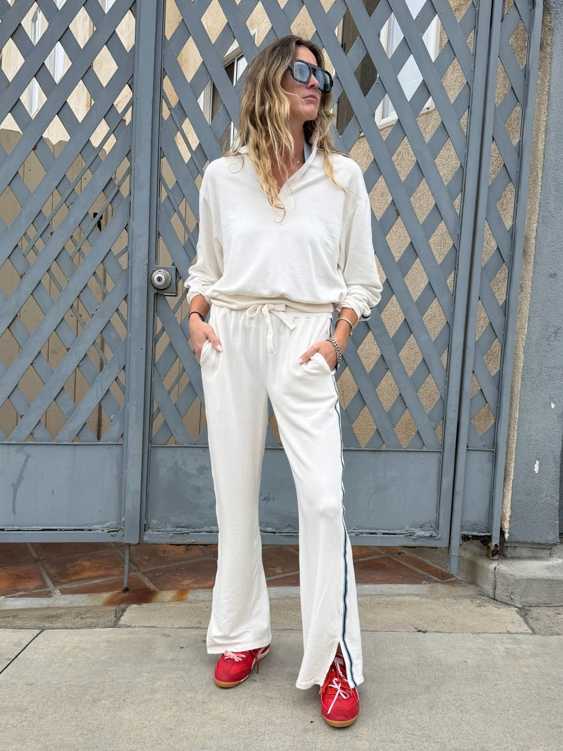 Woman in cream track pants and red shoes stands by a gray gate, facing sideways.