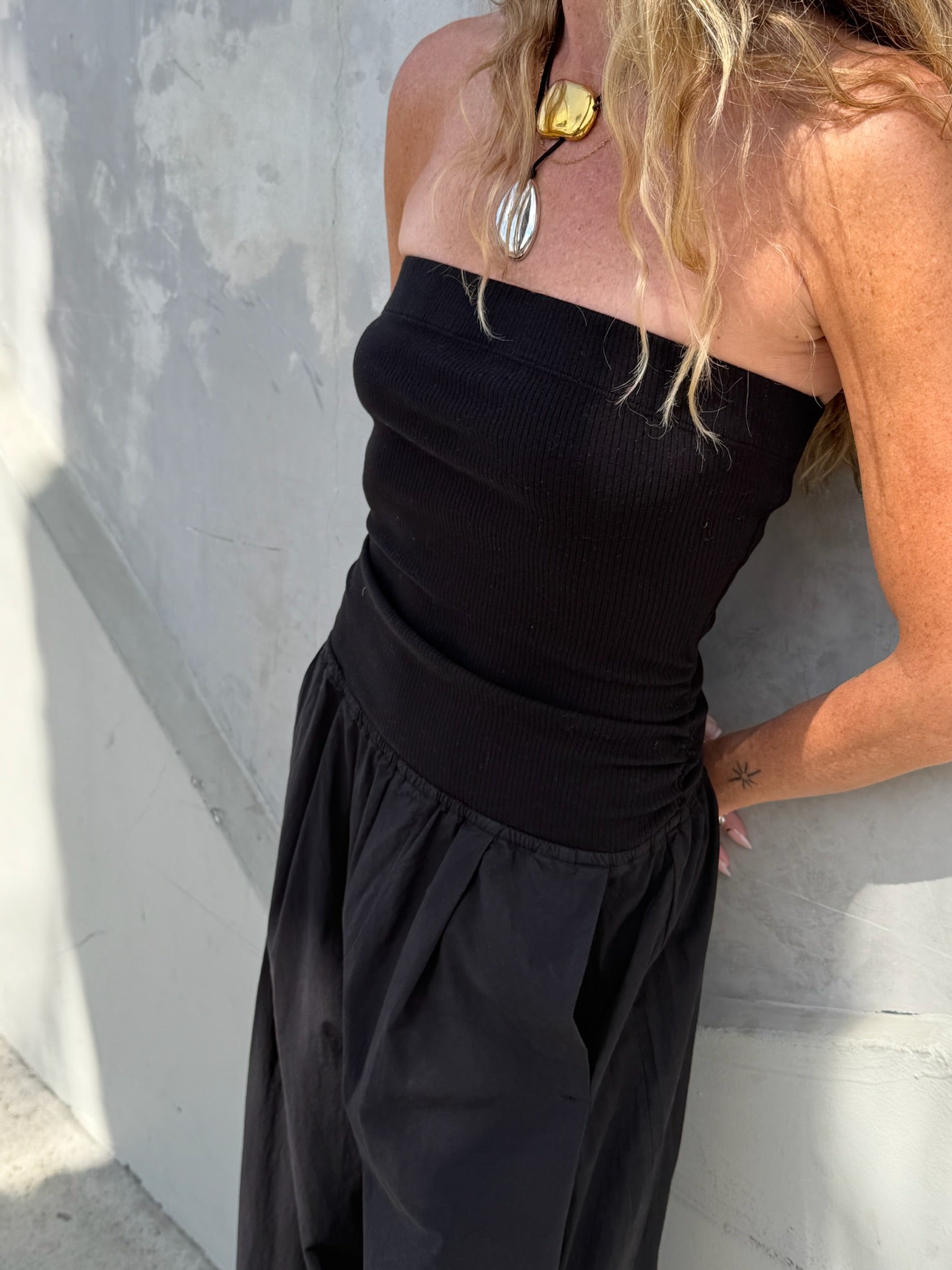 Woman wears a black jumpsuit, gold necklace, and has wavy hair, standing next to a concrete wall.