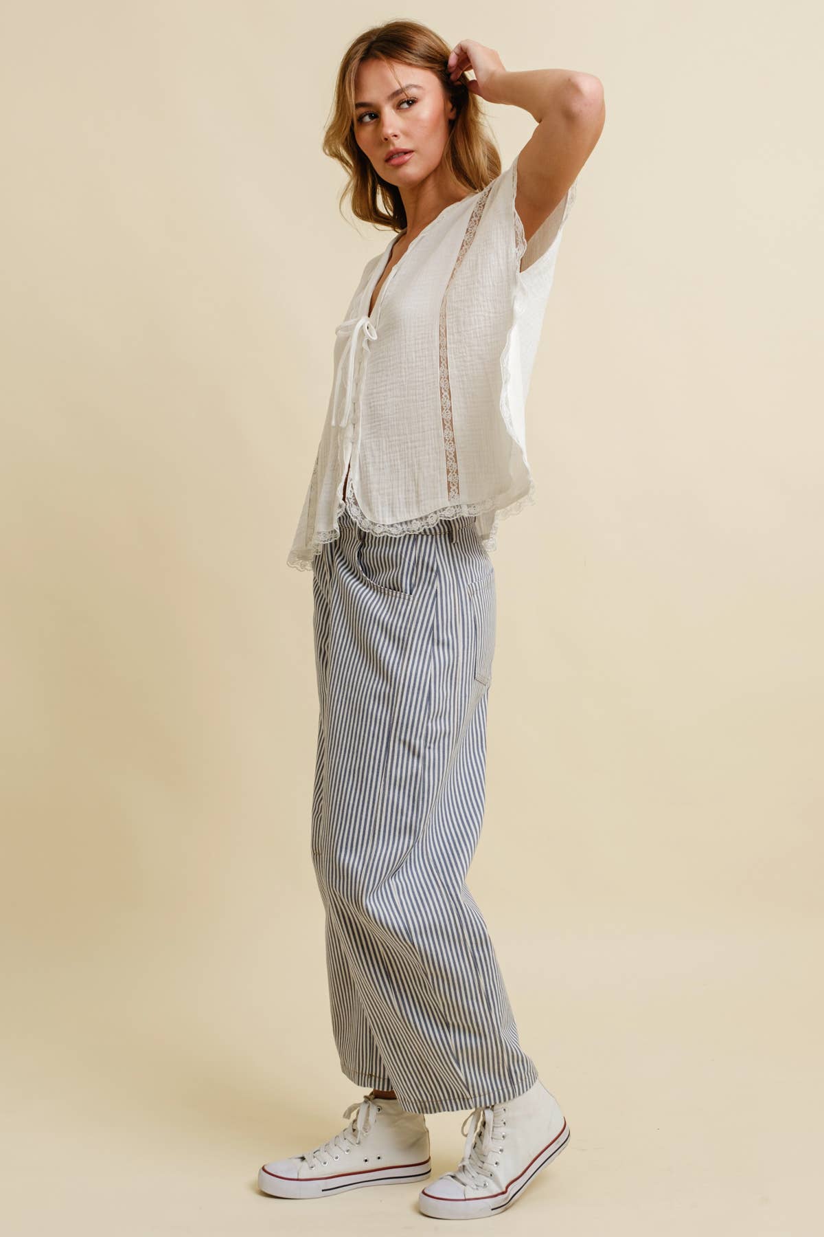 Woman wearing a light top and striped pants, standing sideways, touching her hair, in white high-top sneakers.